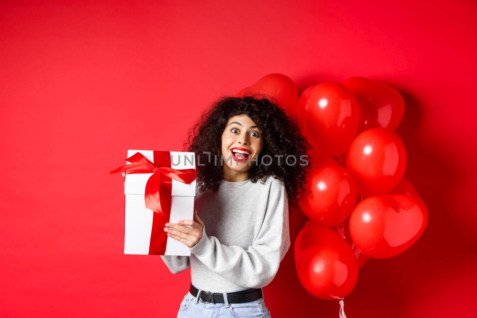 Surprised and happy woman holding valentines day gift from lover, standing near romantic hearts balloons and looking at camera amazed, red background by Benzoix