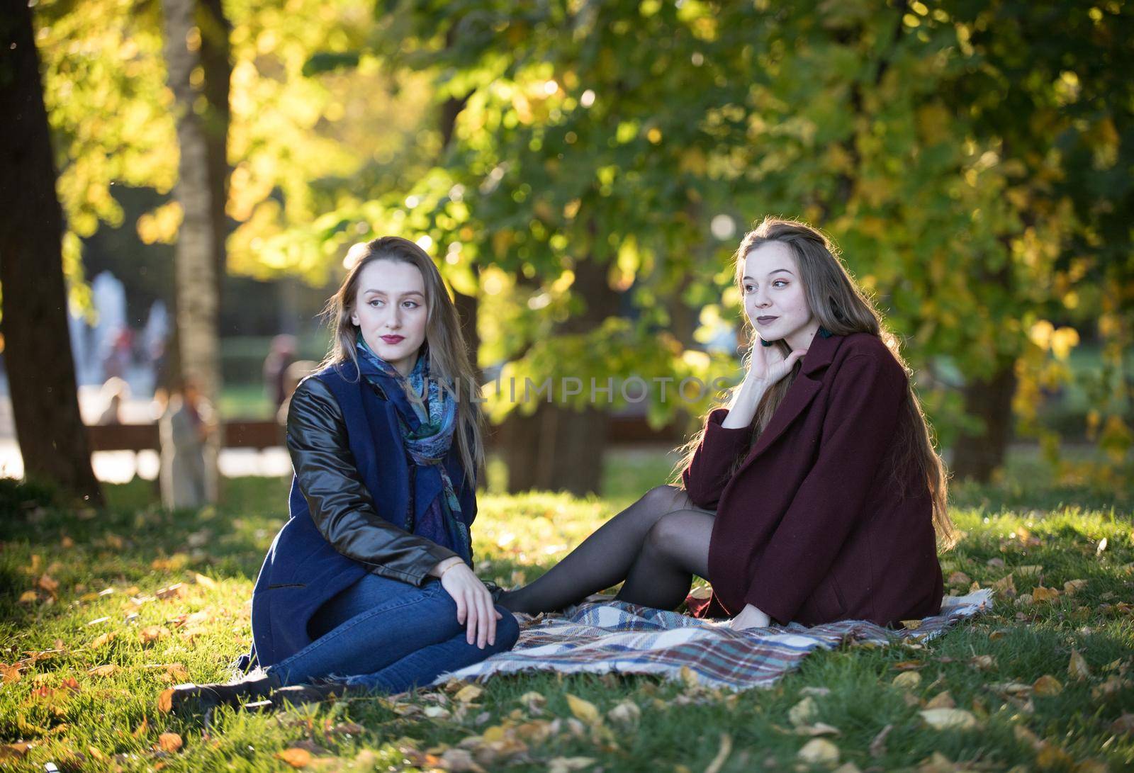 Two beautiful girls in coats sit on a plaid in autumn park in clear weather. Wide shot