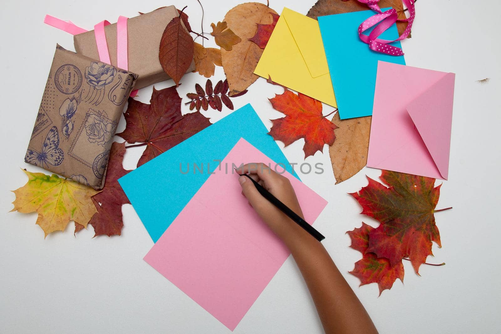 children's hand writes on a pink-colored greeting card Thanksgiving, gifts boxes, envelopes and autumn leaves on a white table, copy space, top view by natashko