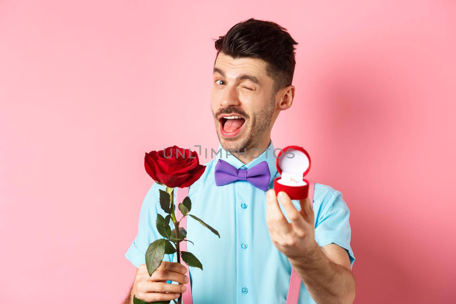 Valentines day. Funny guy making proposal, winking and saying marry me, showing engagement ring with red rose, standing over pink background by Benzoix