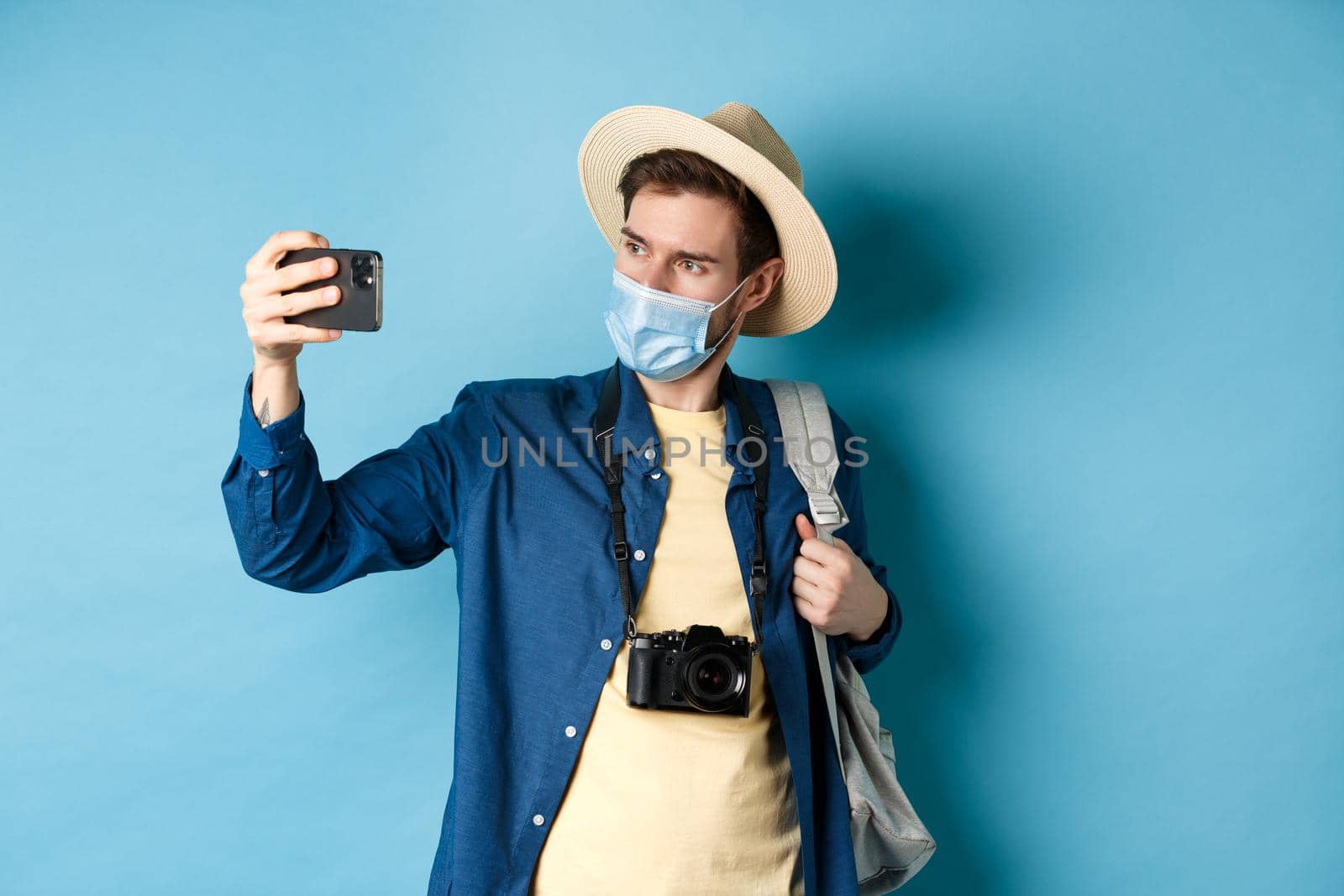 Covid-19, pandemic and travel concept. Male tourist in straw hat and medical mask recording video on smartphone during vacation, taking pics on summer holiday, blue background.