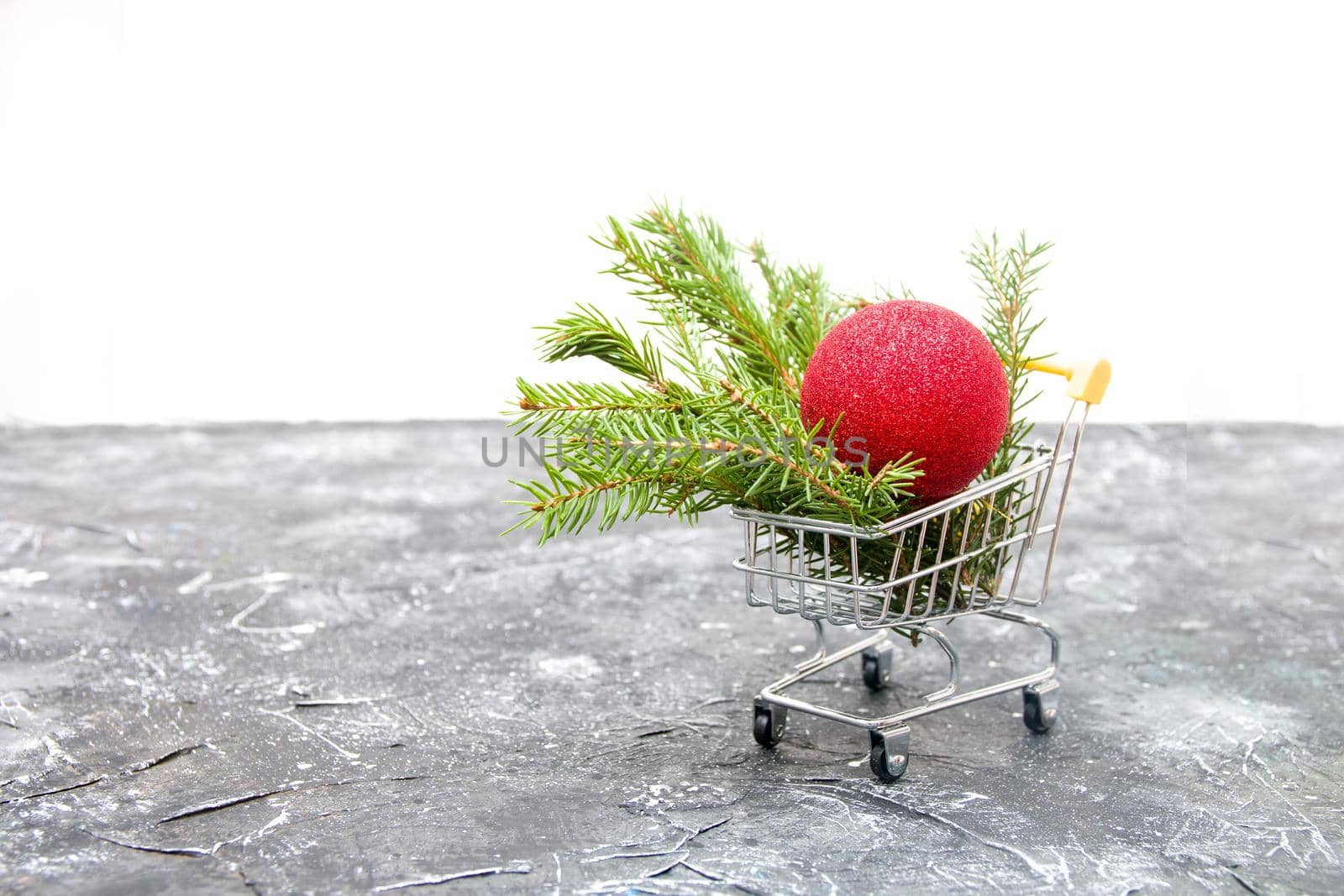 spruce green branch and red shiny Christmas-tree toy ball in a miniature shopping trolley on a black background, copy space