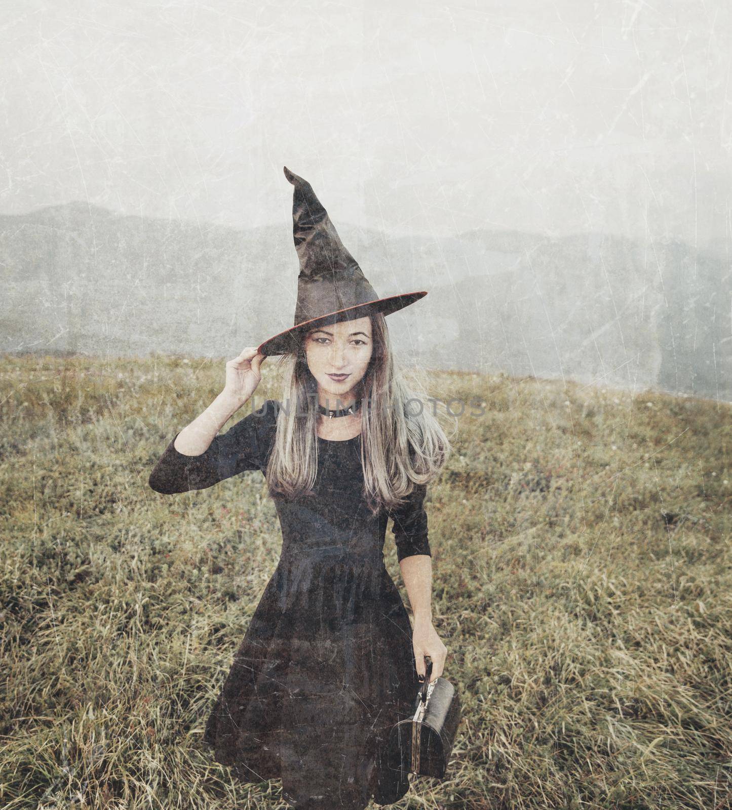 Beautiful witch young woman in hat with handbag walking on meadow outdoor, looking at camera. Theme of Halloween and horror. Vintage image