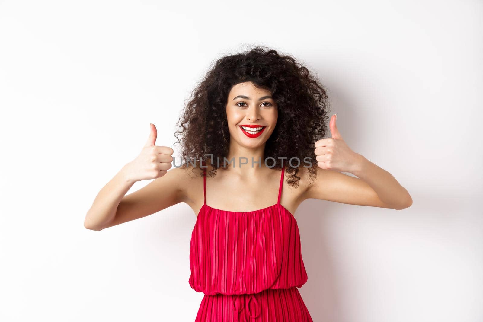 Attractive young woman recommending promo offer, showing thumb up and smiling, like product, standing in festive red dress on white background.