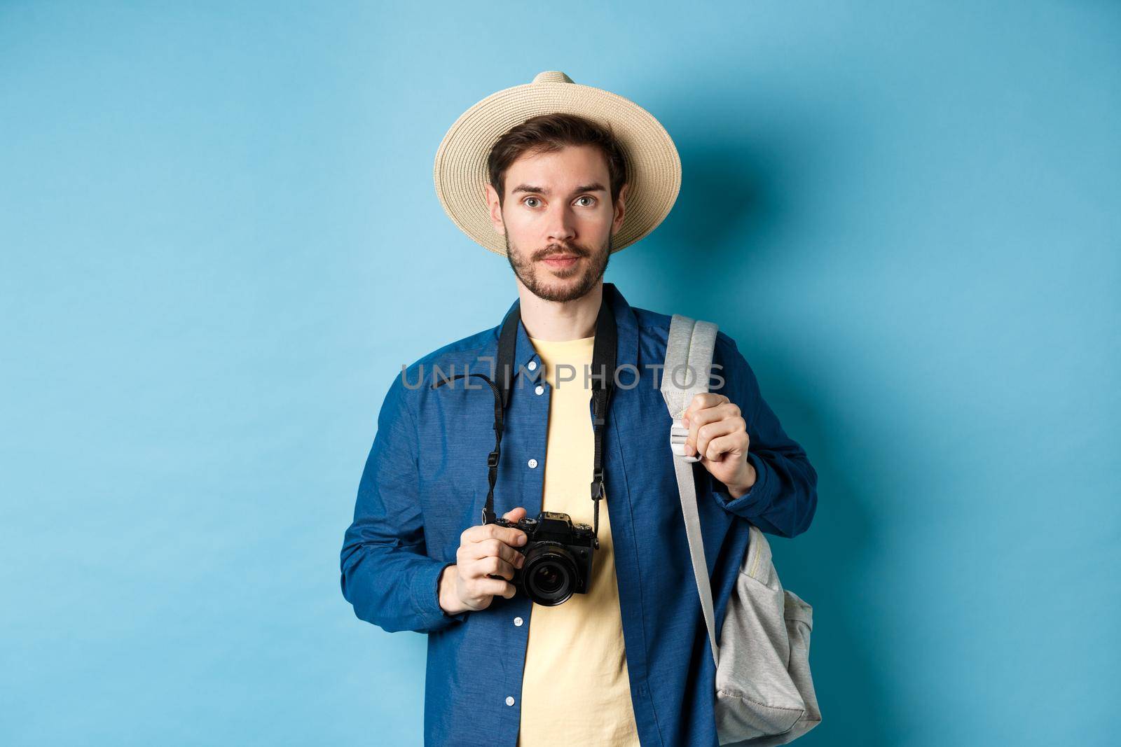 Handsome young tourist in summer hat, backpacking and travelling, holding photo camera, standing on blue background by Benzoix