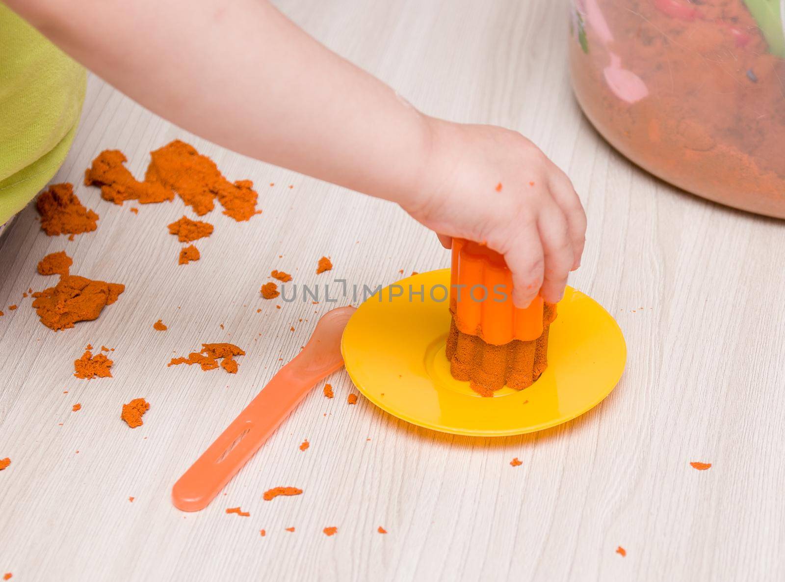 children's hand presses orange sand in a mold for toy muffins and cakes on a beige wooden table by natashko