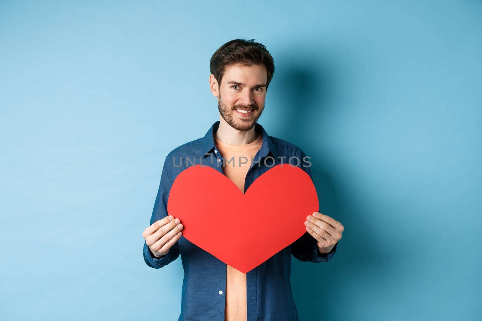 Handsome young man smiling, showing big red heart postcard for Valentines day, looking at camera happy, standing against blue background.