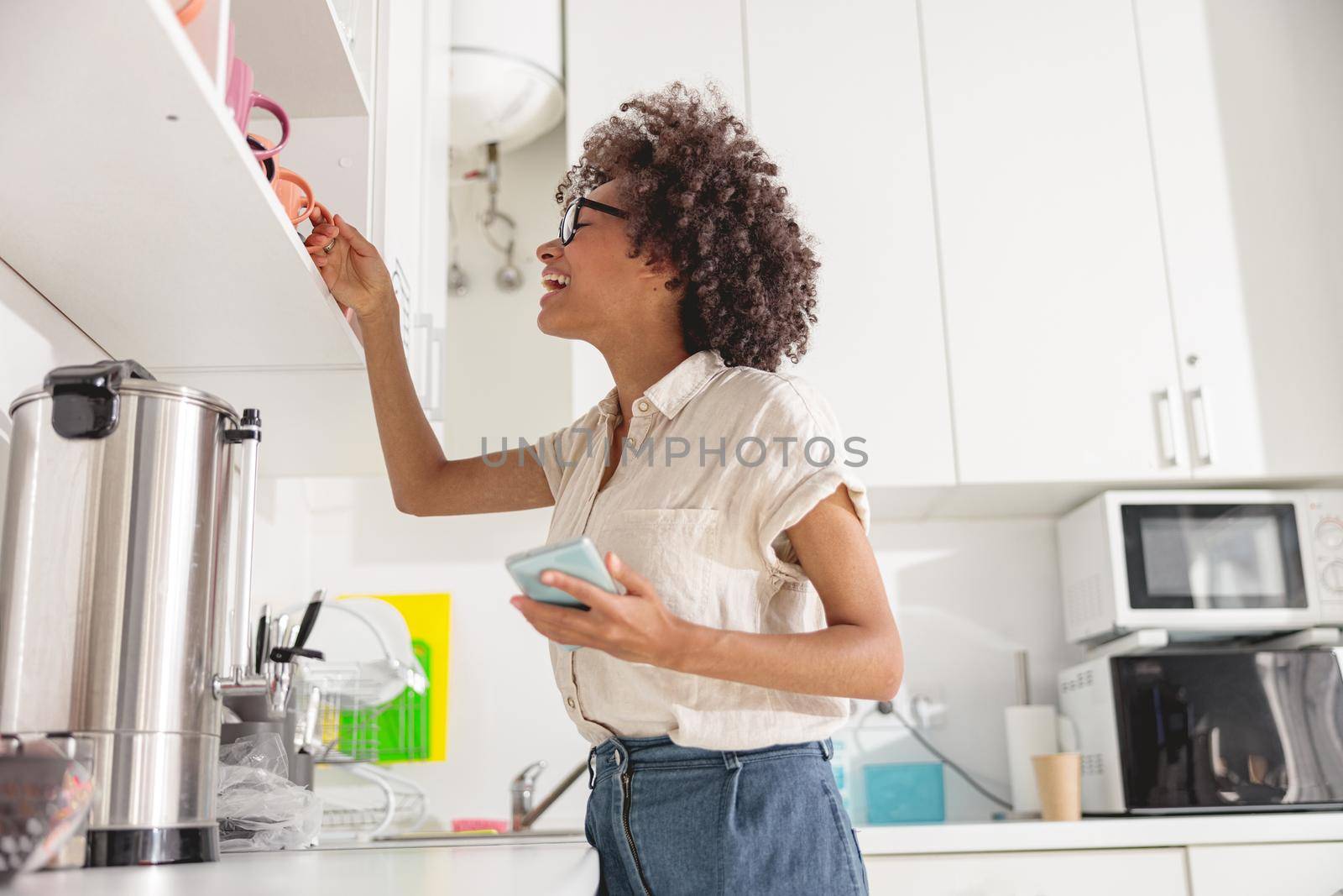 Smiling Afro American lady holding mobile phone while going to make coffee on office kitchen