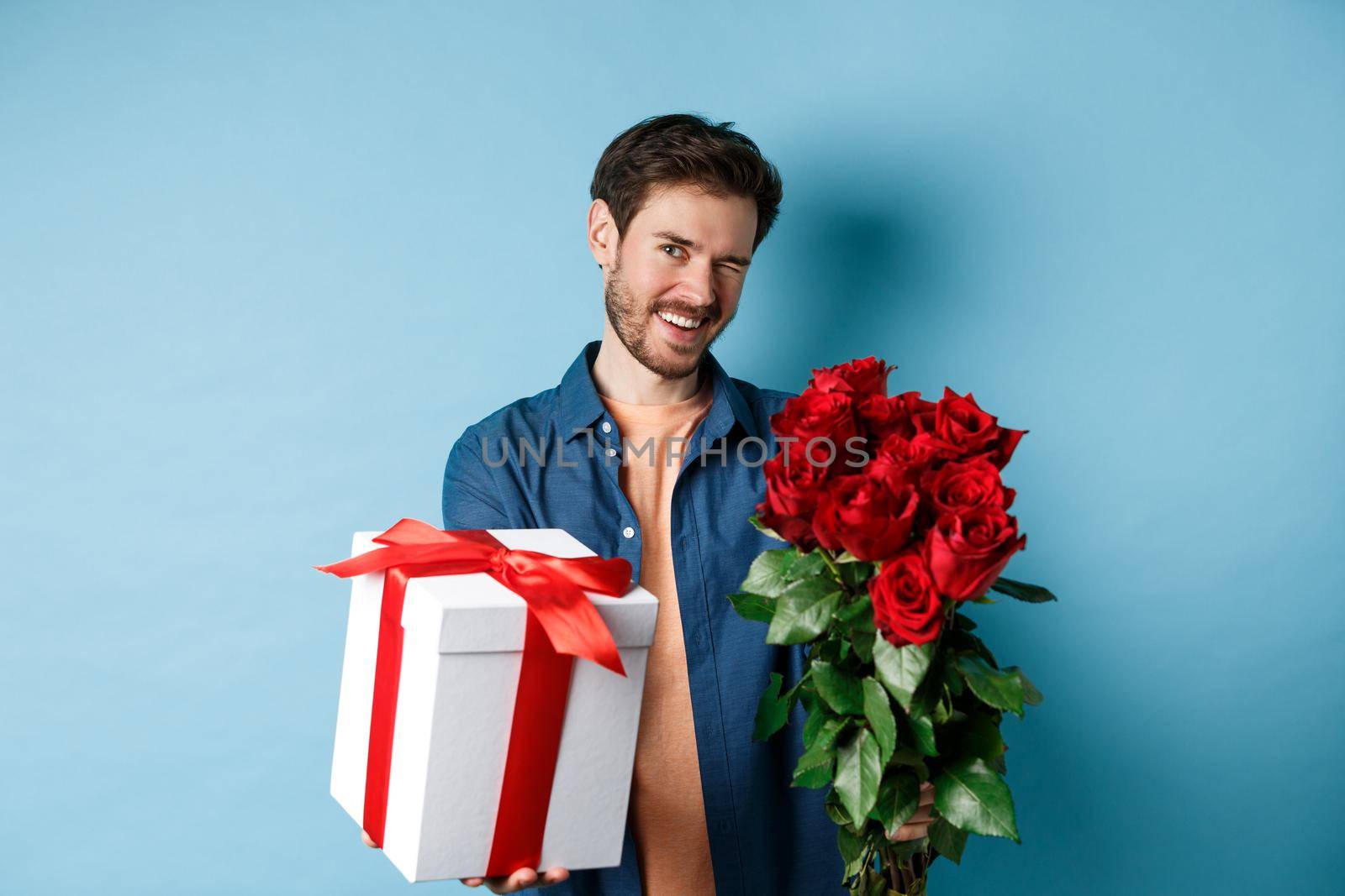 Love and Valentines day concept. Happy guy winking and smiling at camera, presenting a gift and bouquet of flowers to girlfriend, standing over blue background.