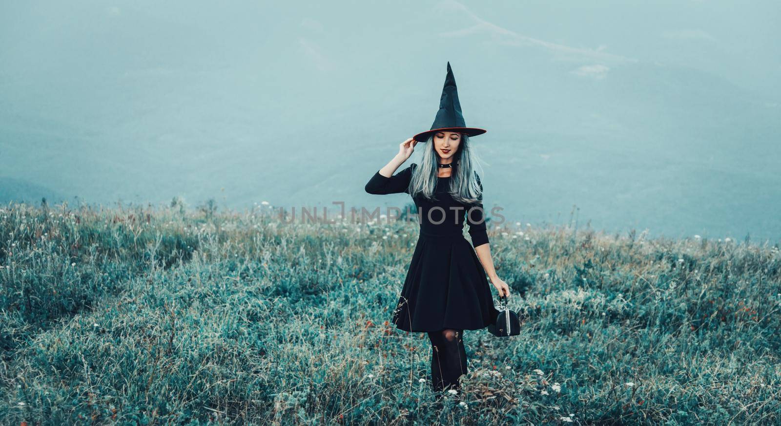 Beautiful fashionable young witch in black dress and hat with small bag walking on autumn meadow. Halloween theme