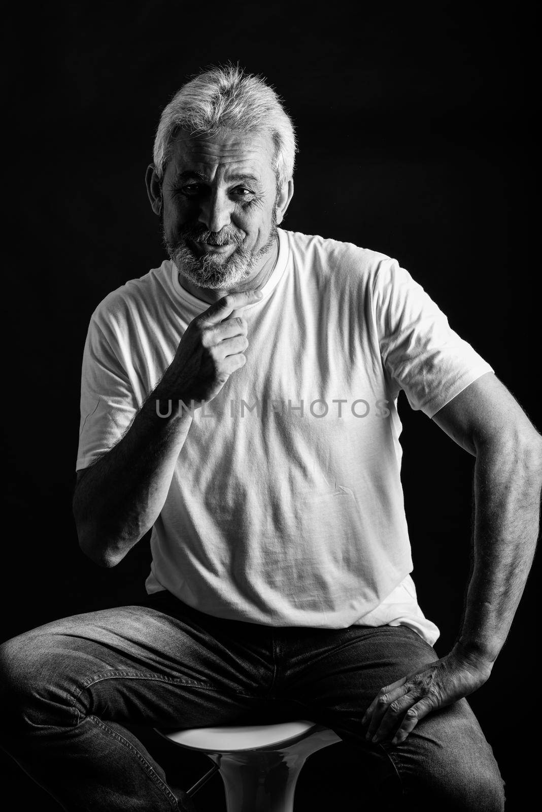 Mature man smiling looking at camera. Senior male with white hair and beard laughing wearing casual clothes isolated on black background. Studio shot in black and white.