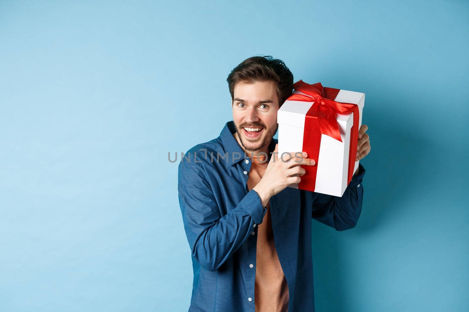 Valentines day. Happy young man got present on special holiday, trying guess what inside gift box and smiling, standing over blue background.