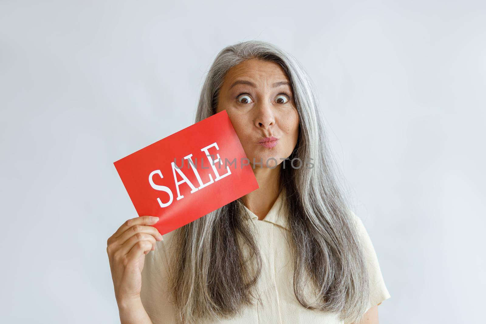 Funny middle aged Asian woman shows red Sale sign grimacing on light background by Yaroslav_astakhov