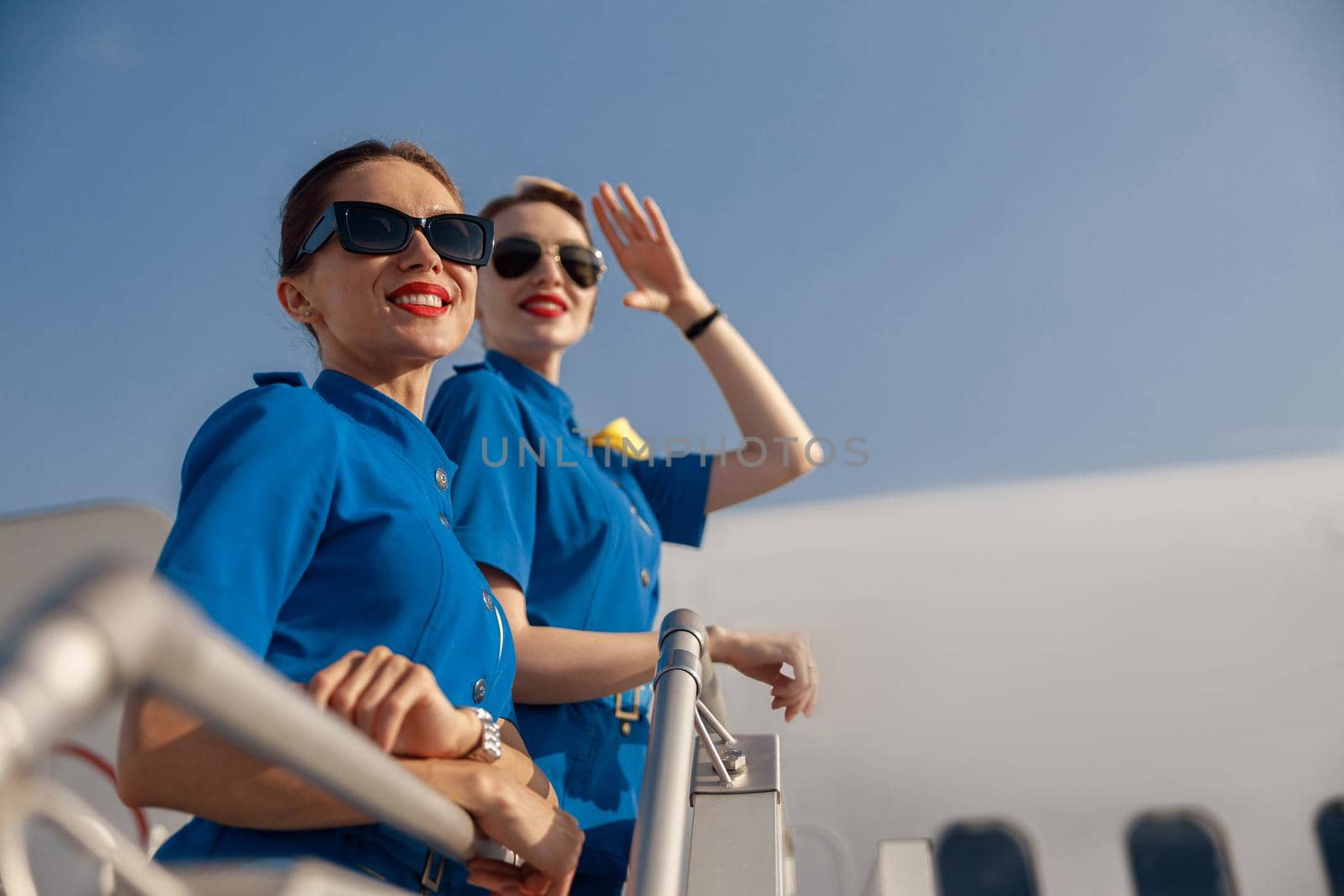 Portrait of two cheerful air stewardesses in blue uniform and sunglasses smiling away, standing together on airstair on a sunny day. Aircrew, occupation concept