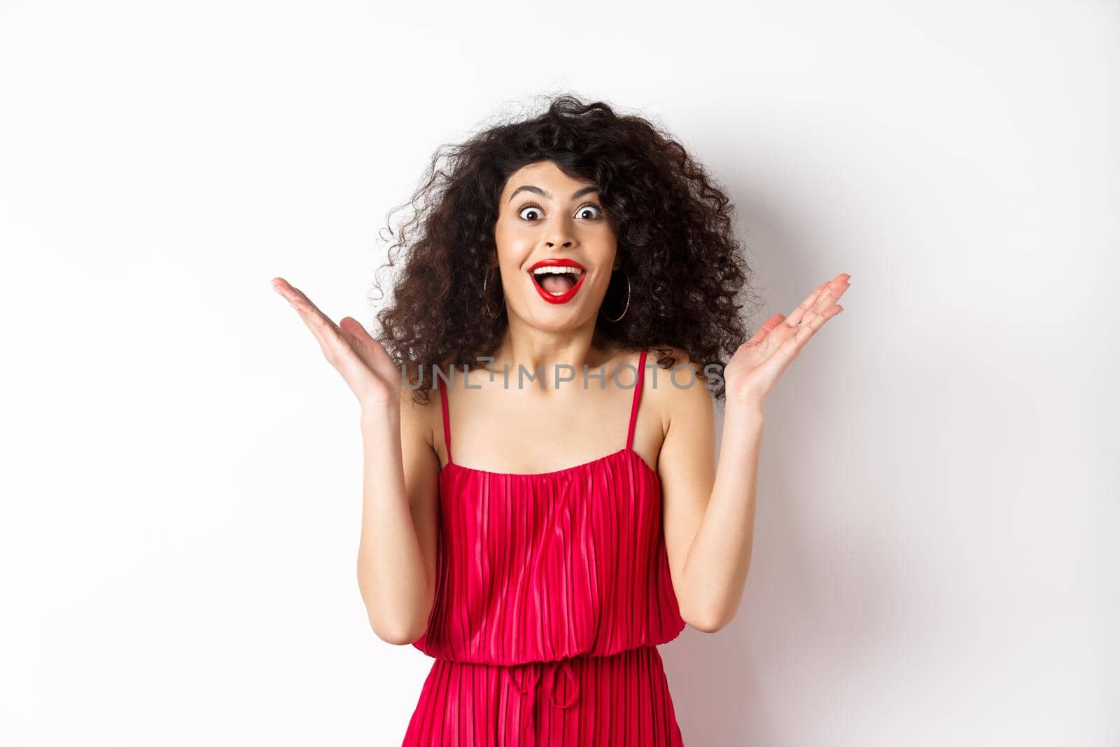 Surprised happy woman reacting to big news, looking cheerful with hands spread sidewas, standing in red dress on white background.