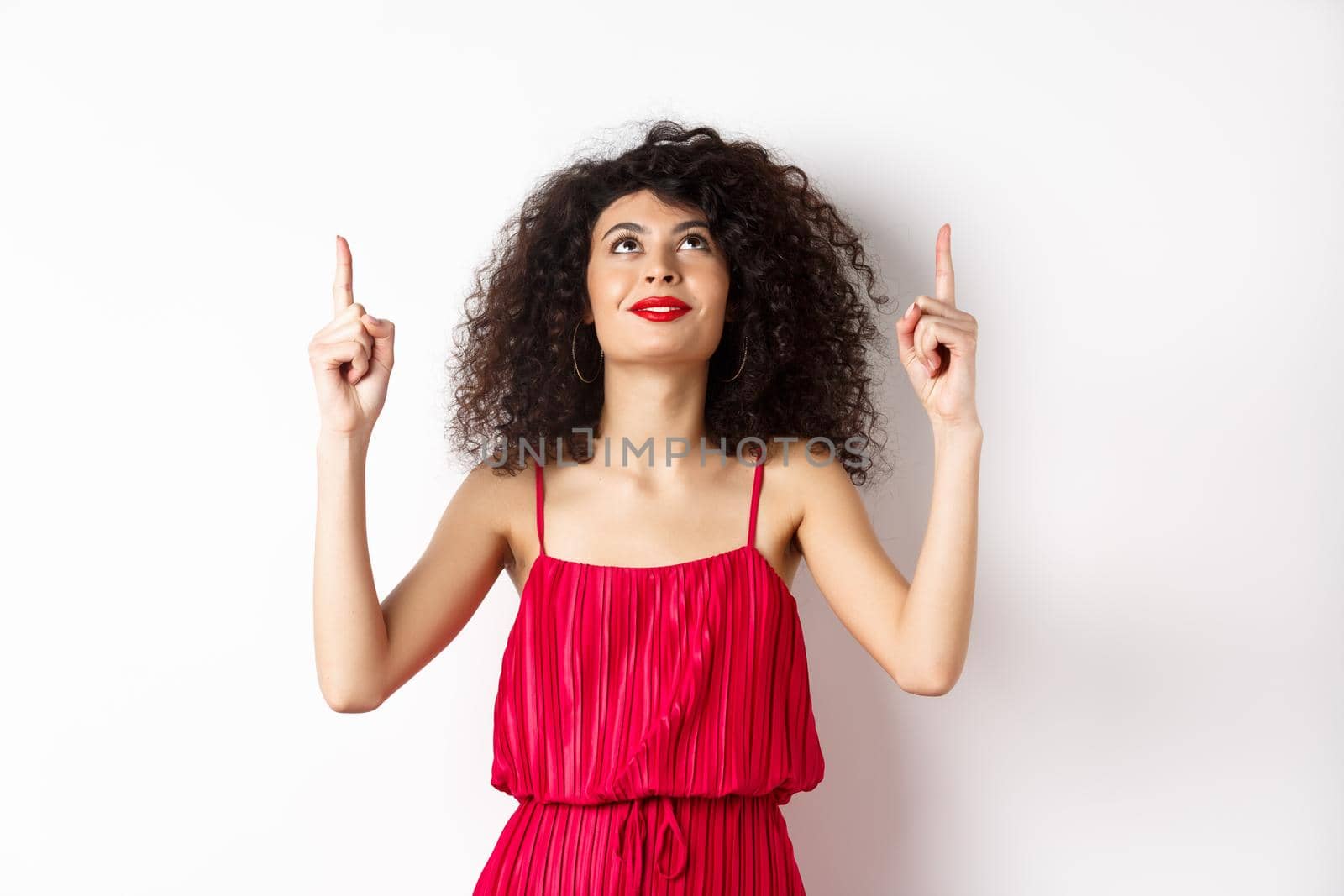 Dreamy young woman in trendy red dress, looking up and pointing at logo, smiling happy, standing over white background. Copy space