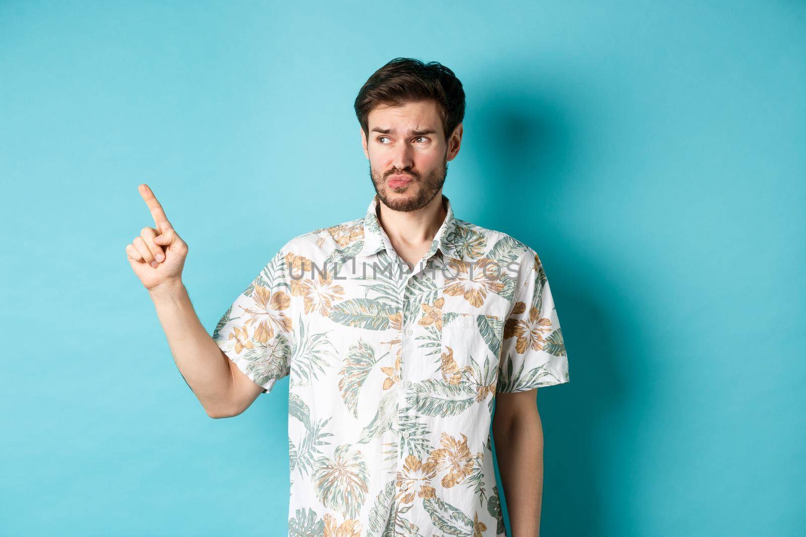 Displeased tourist in hawaiian shirt, looking and pointing left disappointed, complaining something bad, standing on blue background.