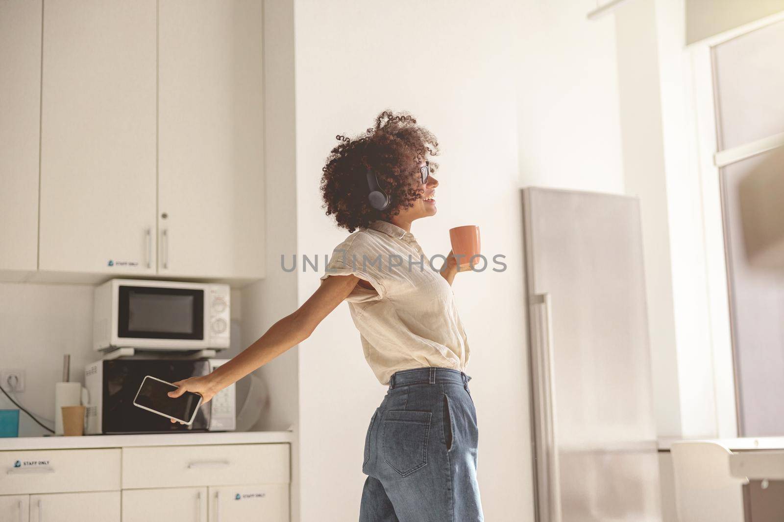 Happy Afro American lady wearing wireless headphones and dancing in the kitchen while holding the mobile phone