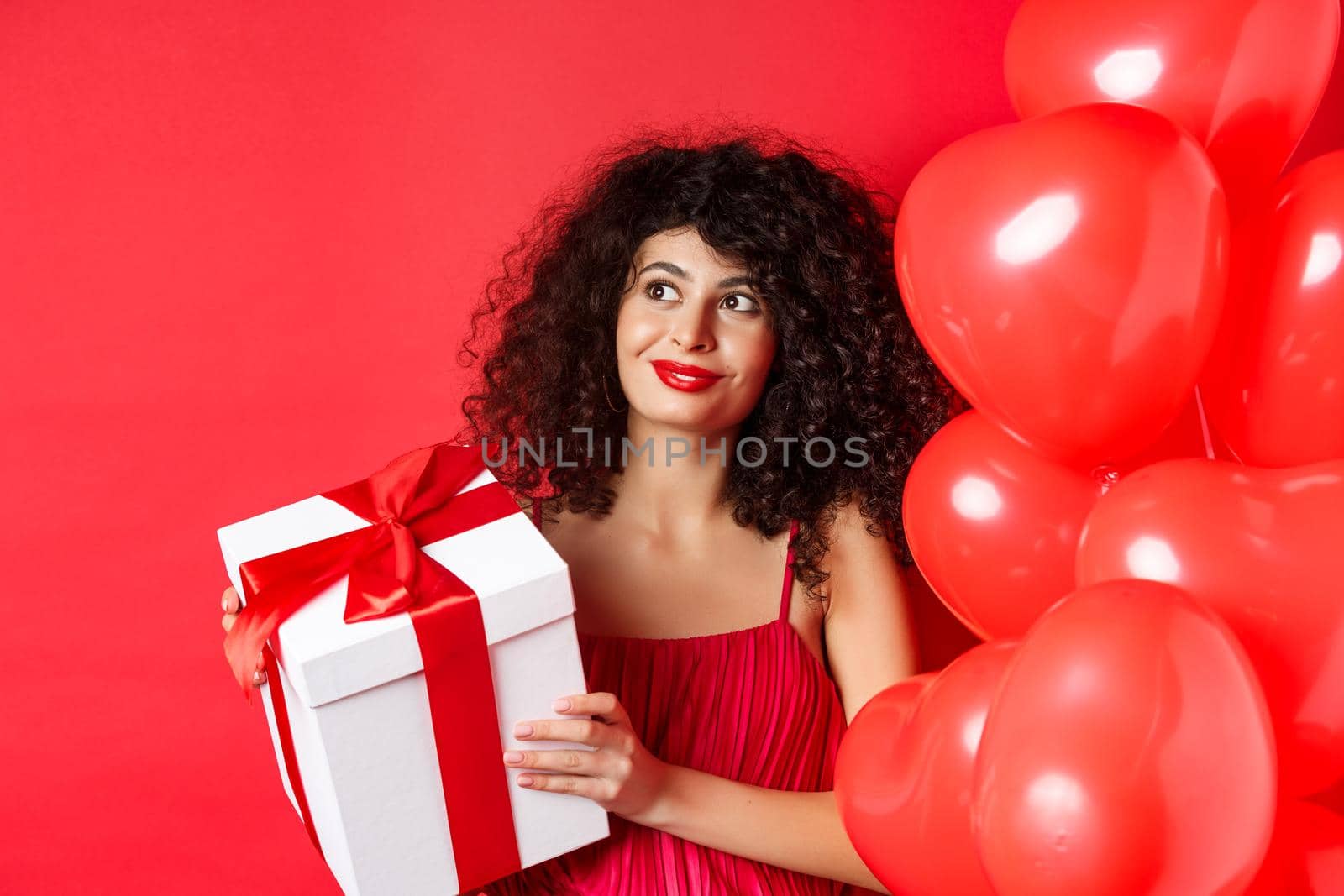 Gorgeous woman with makeup and evening dress, receive surprise gift and smiling, standing on red background near Valentines day romantic balloons by Benzoix