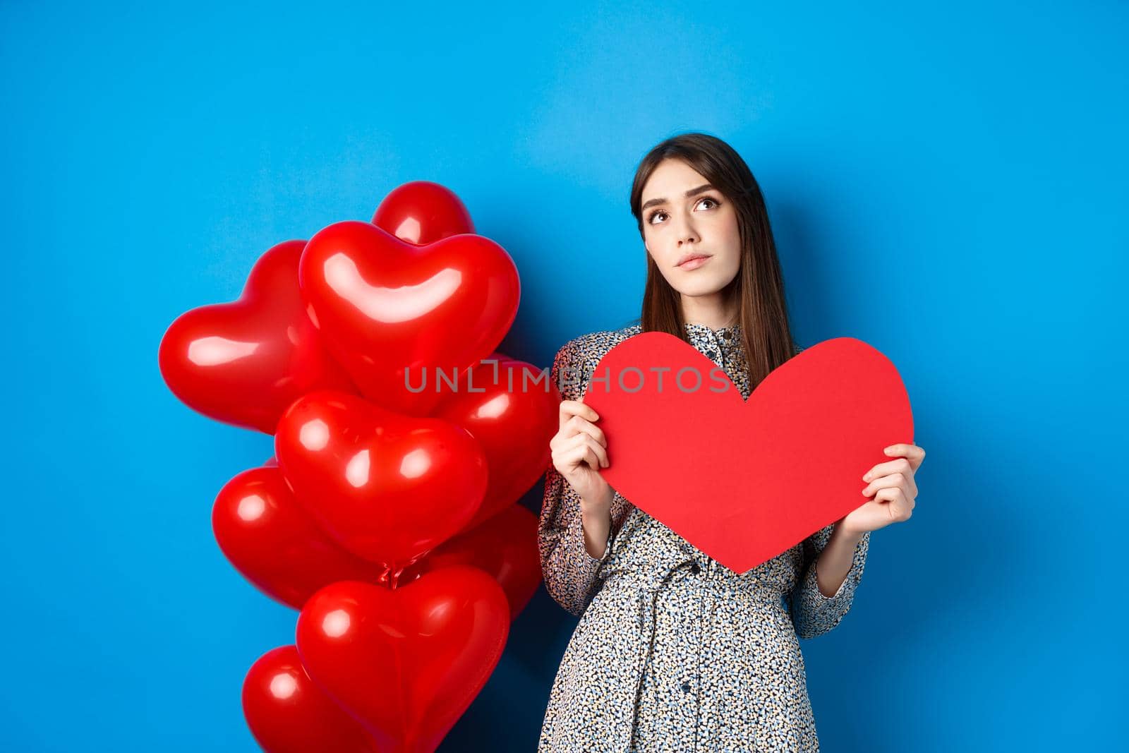 Valentines day. Dreamy pretty lady in dress, holding big red heart cutout and searching for true love, looking up pensive, standing near holiday balloons on blue background by Benzoix