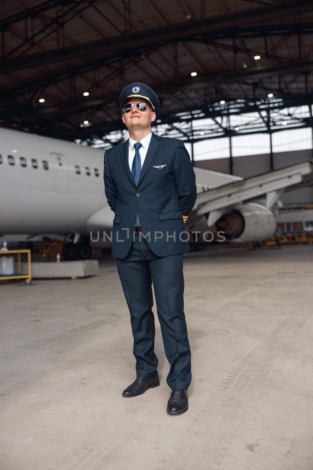 Full length shot of proud pilot in uniform and aviator sunglasses smiling away, standing in front of big passenger airplane in airport hangar. Aircraft, occupation, transportation concept