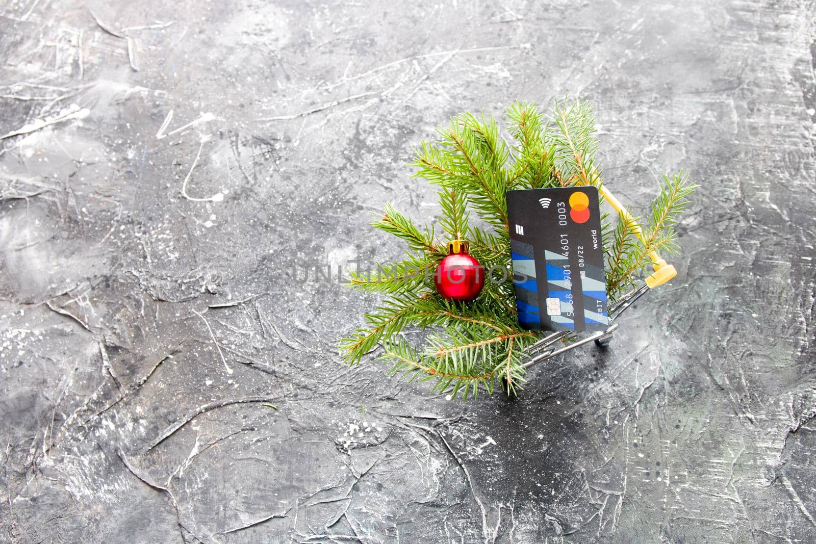 spruce green branch and red shiny Christmas-tree toy ball in a miniature shopping trolley on a black background, copy space by natashko