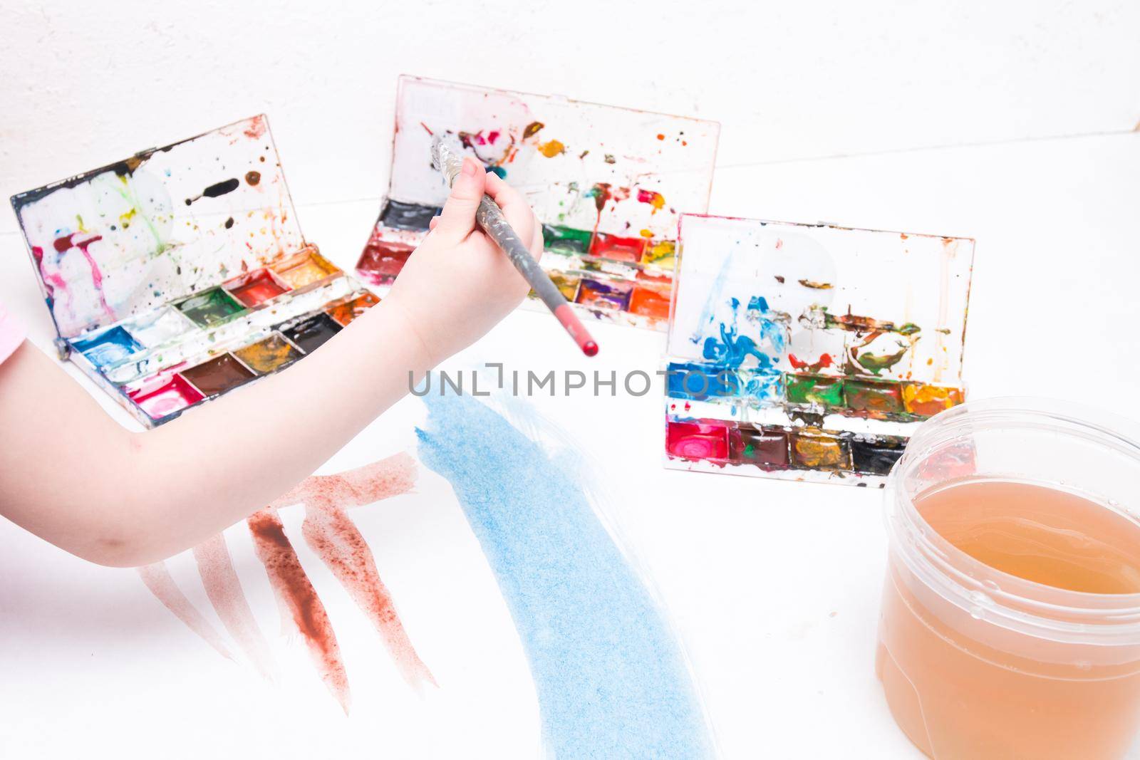 the child paints with a brush watercolors on a white sheet, a table, a copy space, several packages of watercolor, a large art brush in his hand, a can of water