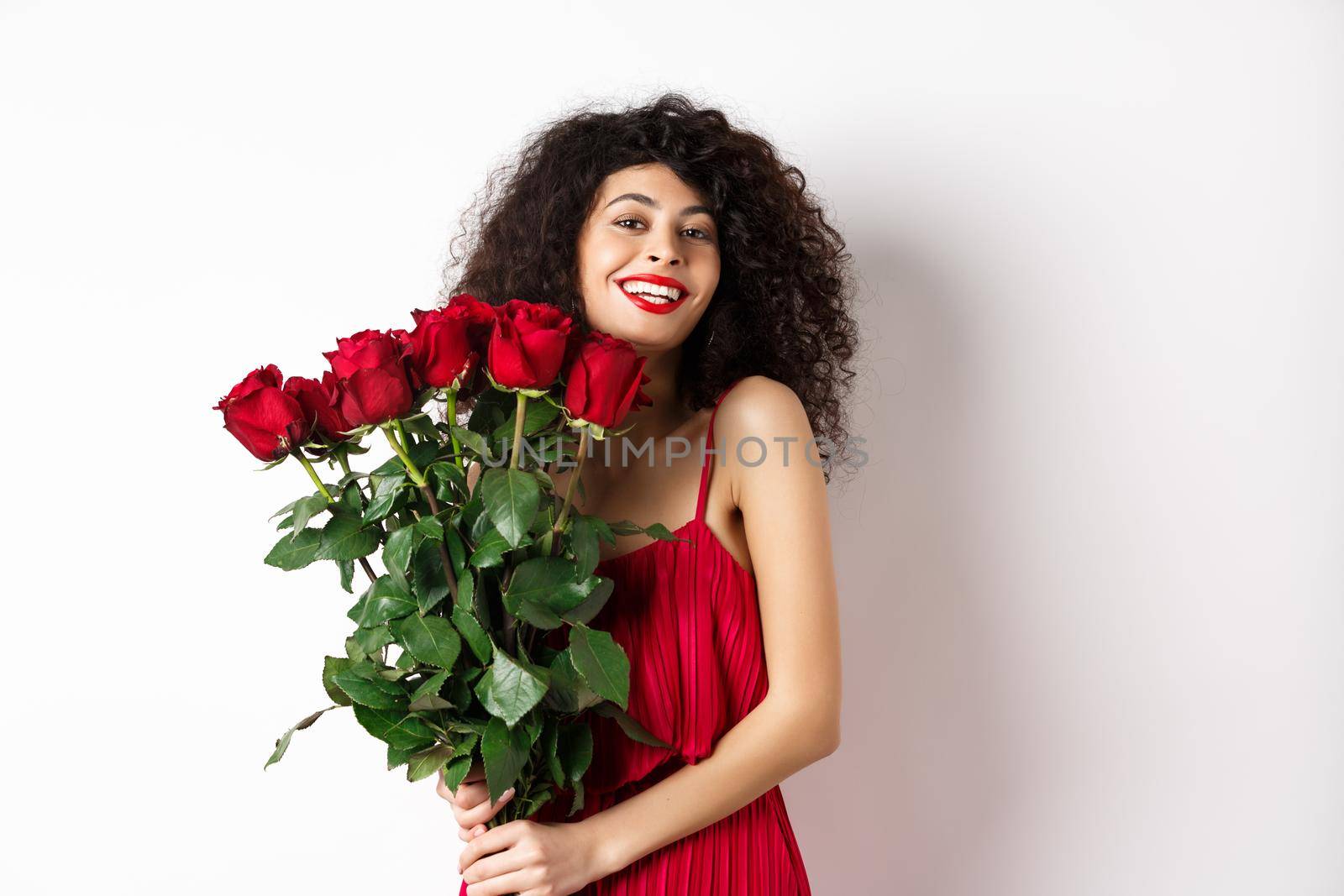 Romantic lady with curly hair and fashionable dress, holding bouquet of red roses and smiling, standing happy on white background by Benzoix