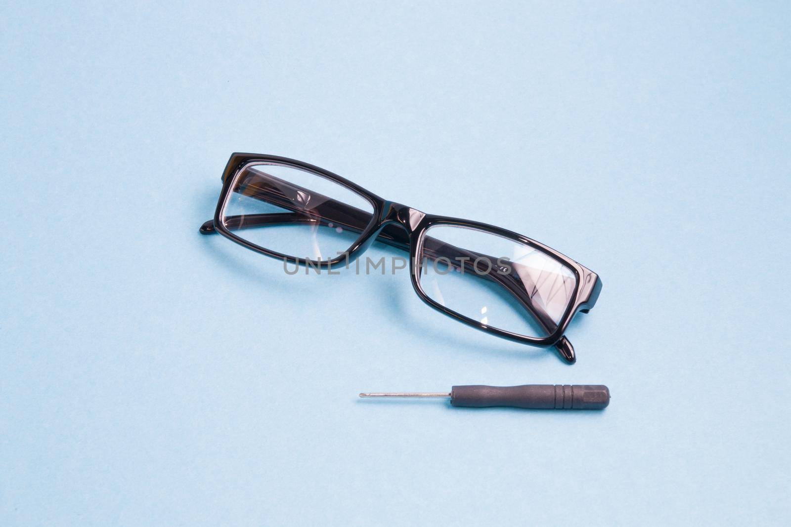 glasses in a black plastic frame and a small screwdriver are on a light blue background, broken glasses are under repair