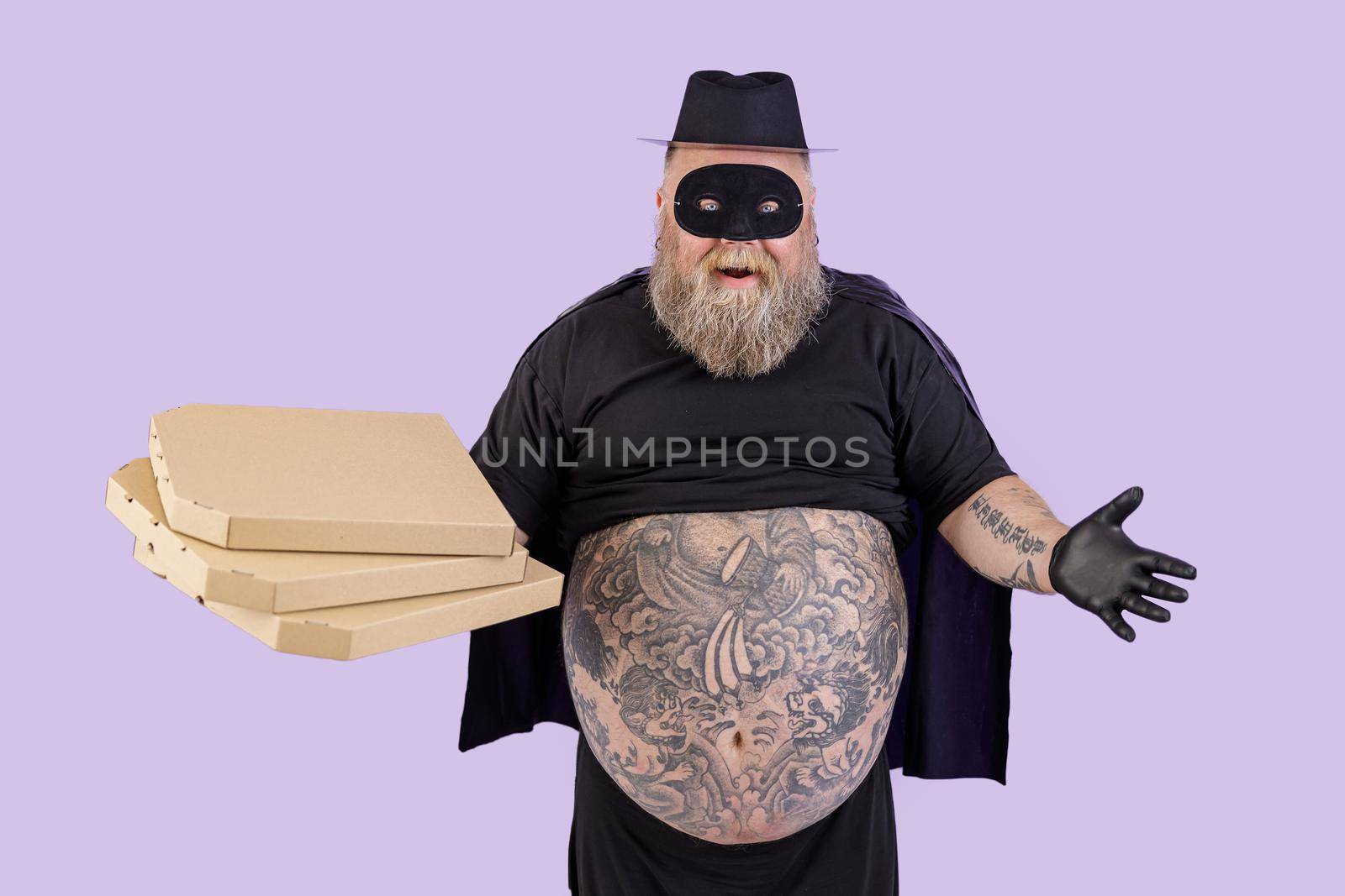 Positive emotional mature fat man in Zorro costume with large tattooed tummy holds cardboard boxes of pizza standing on purple background in studio