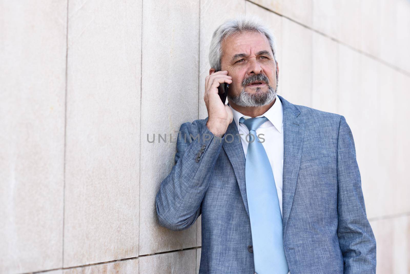 Portrait of a senior businessman talking with a smart phone outside of modern office building. Successful business man wearing blue suit and tie in urban background. Male in formal clothes