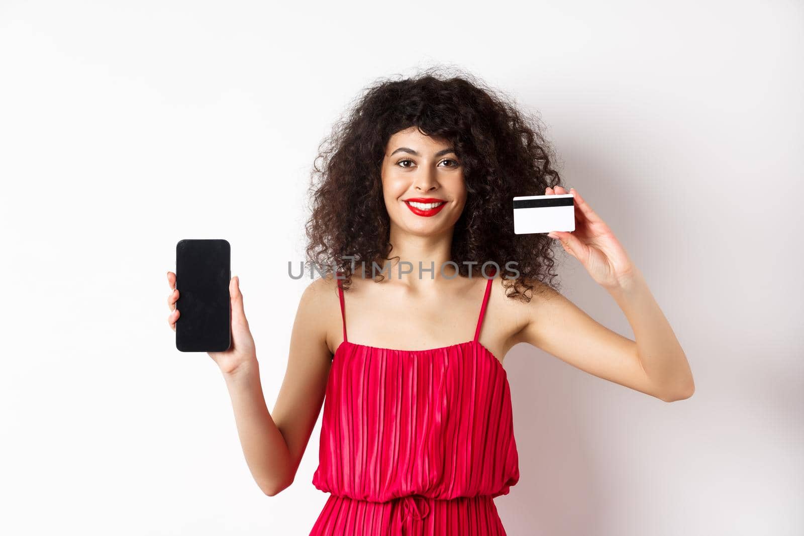 Online shopping concept. Elegant female model with curly hair, wearing red dress, showing plastic credit card with empty mobile phone screen, standing on white background by Benzoix