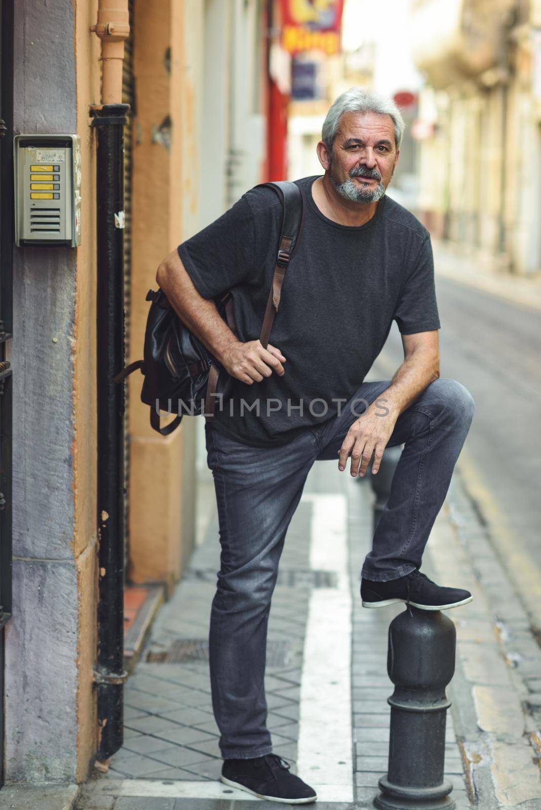 Mature tourist man in urban background. Mature male with white hair and beard wearing casual clothes and travel backpack