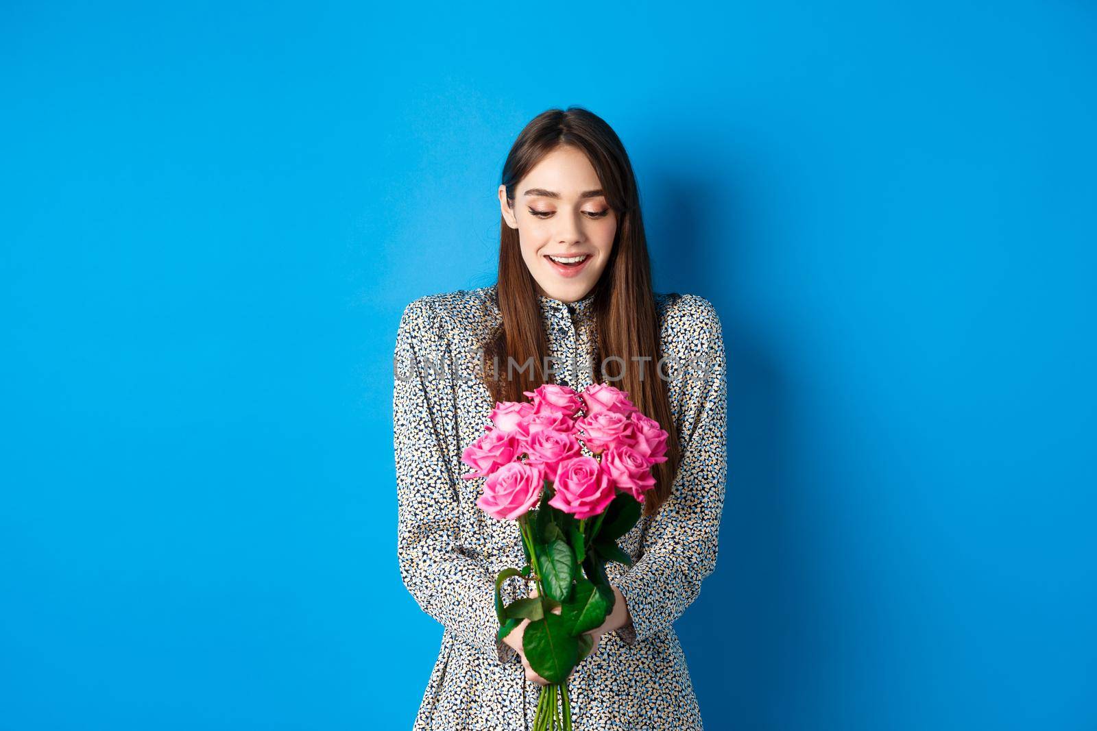 Valentines day concept. Happy attractive woman receive surprise flowers, looking thankful at bouquet of pink roses, standing on blue background.