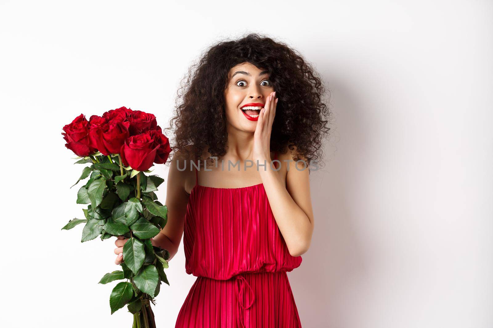 Excited curly woman in red dress, receive bouquet of roses and look surprise, rejoicing from romantic gift, standing on white background by Benzoix