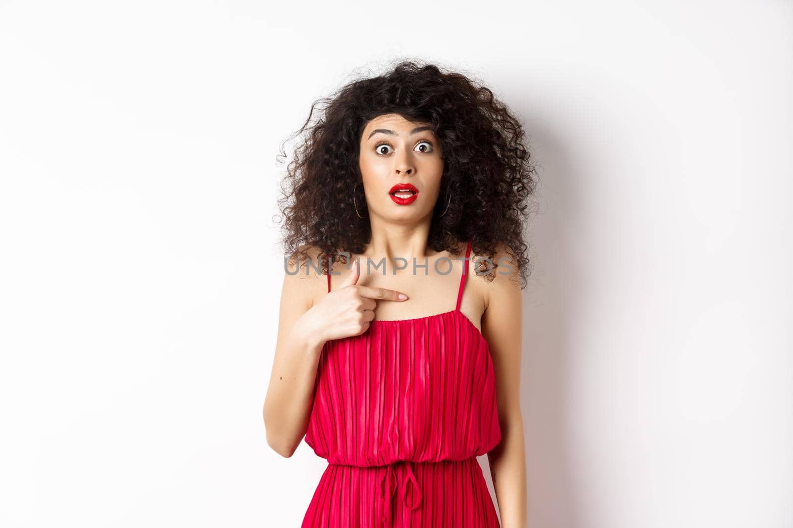 Surprised young woman in red dress pointing at herself, being confused and shocked, standing on white background by Benzoix