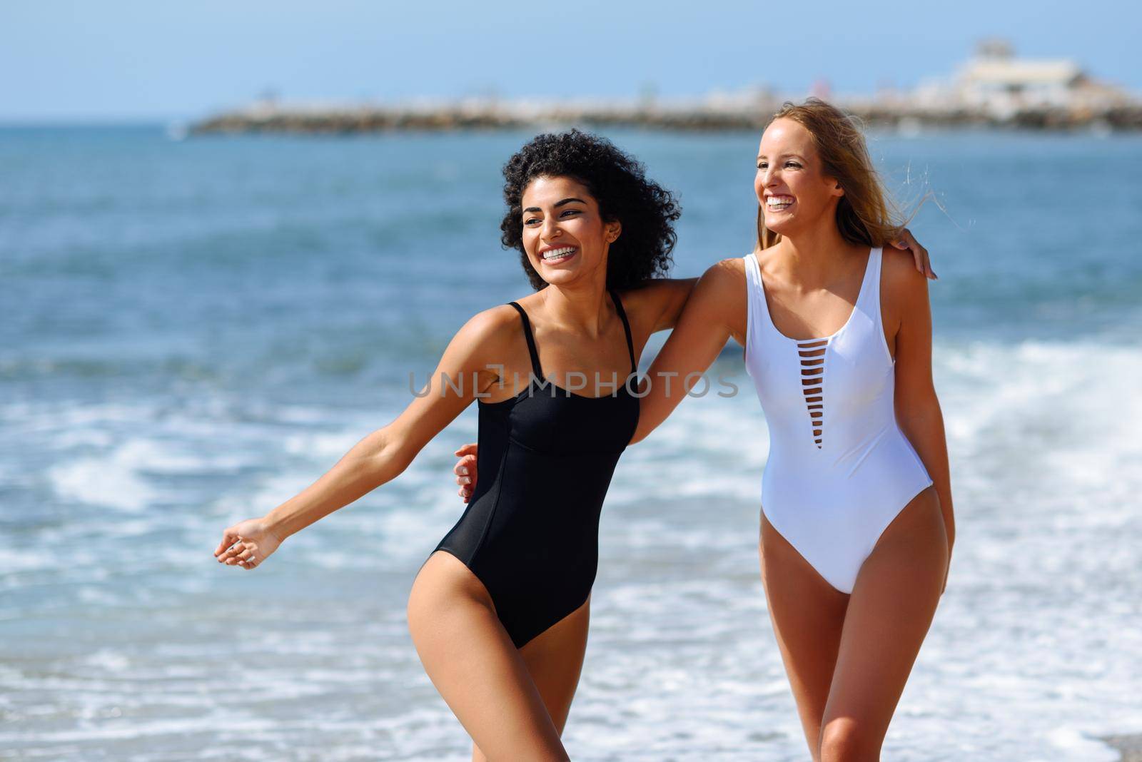 Two young women with beautiful bodies in swimwear on a tropical beach. Funny caucasian and arabic females wearing black and white swimsuits walking along the shore.