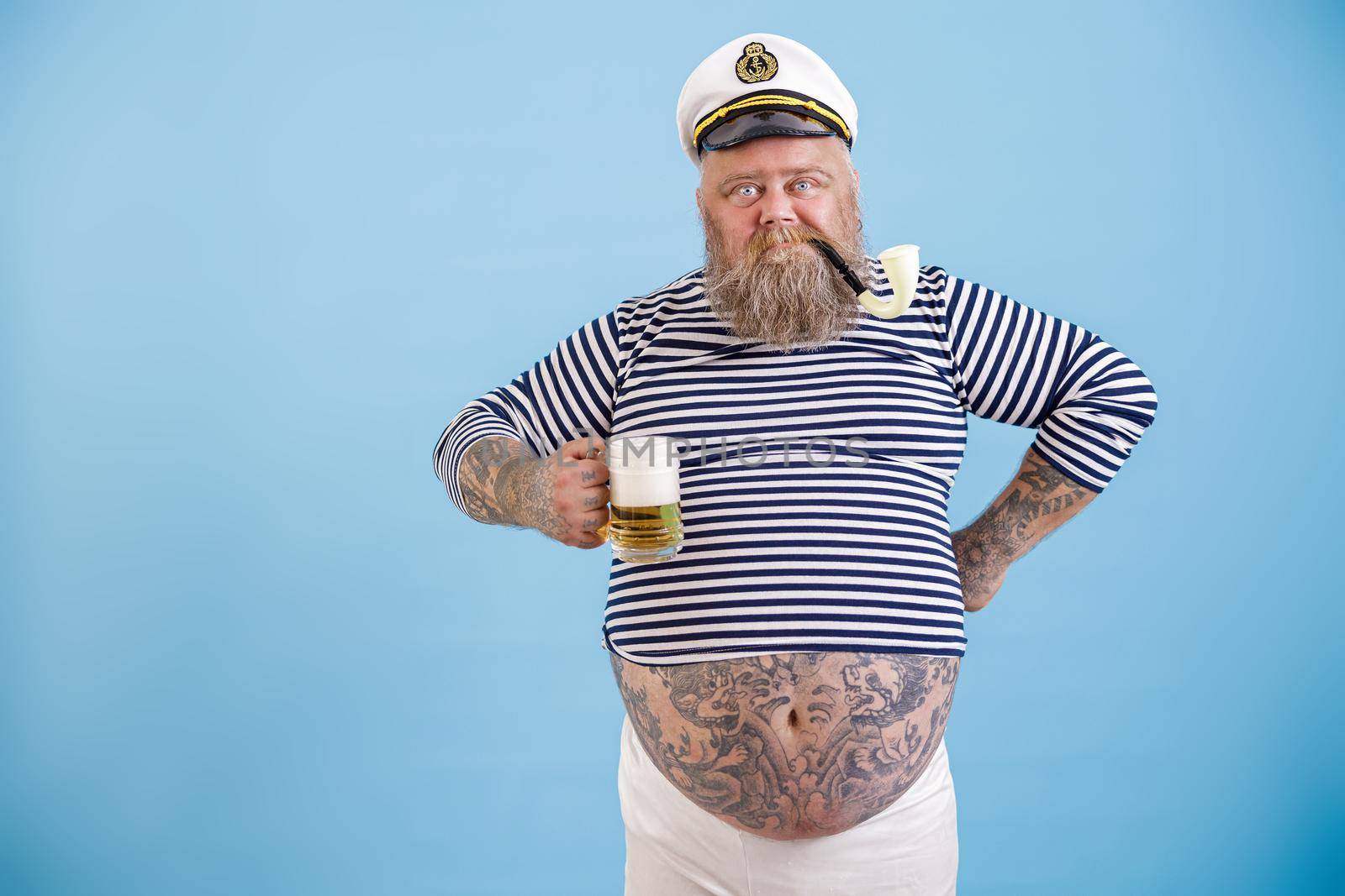 Dashing fat man with long beard in sailor costume with smoking pipe holds glass mug of foamy beer on light blue background in studio