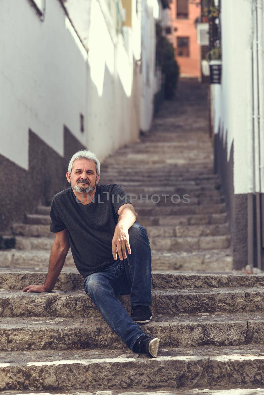 Portrait of a mature man sitting on steps in the street. Senior male with white hair and beard wearing casual clothes in urban background.