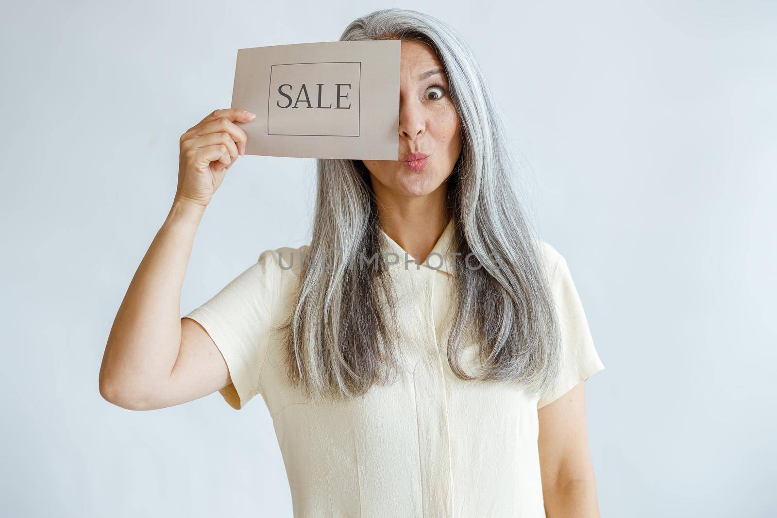Funny mature woman with loose hoary hair covers eye with Sale sign standing on light grey background in studio