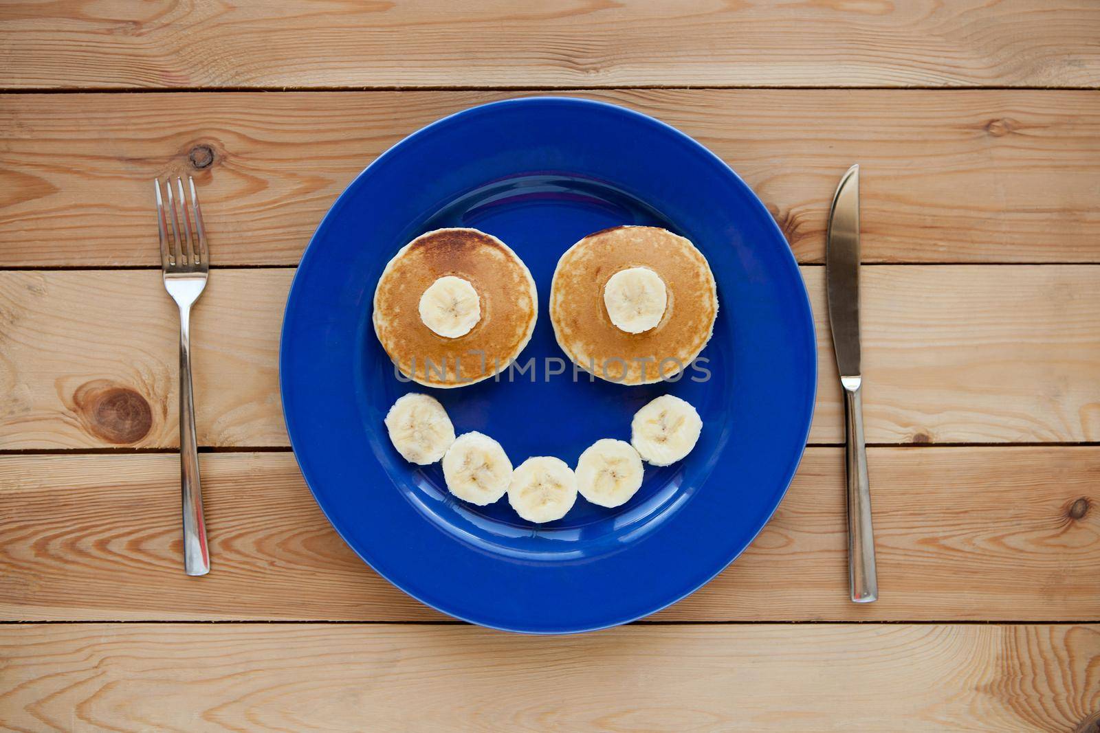 Pancakes with smile on blue plate and wooden background. banana fruit smiling breakfast - fun food for kids