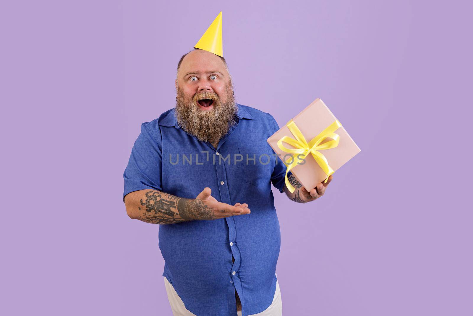 Joyful bearded man with overweight holds gift box with bow posing on purple background by Yaroslav_astakhov