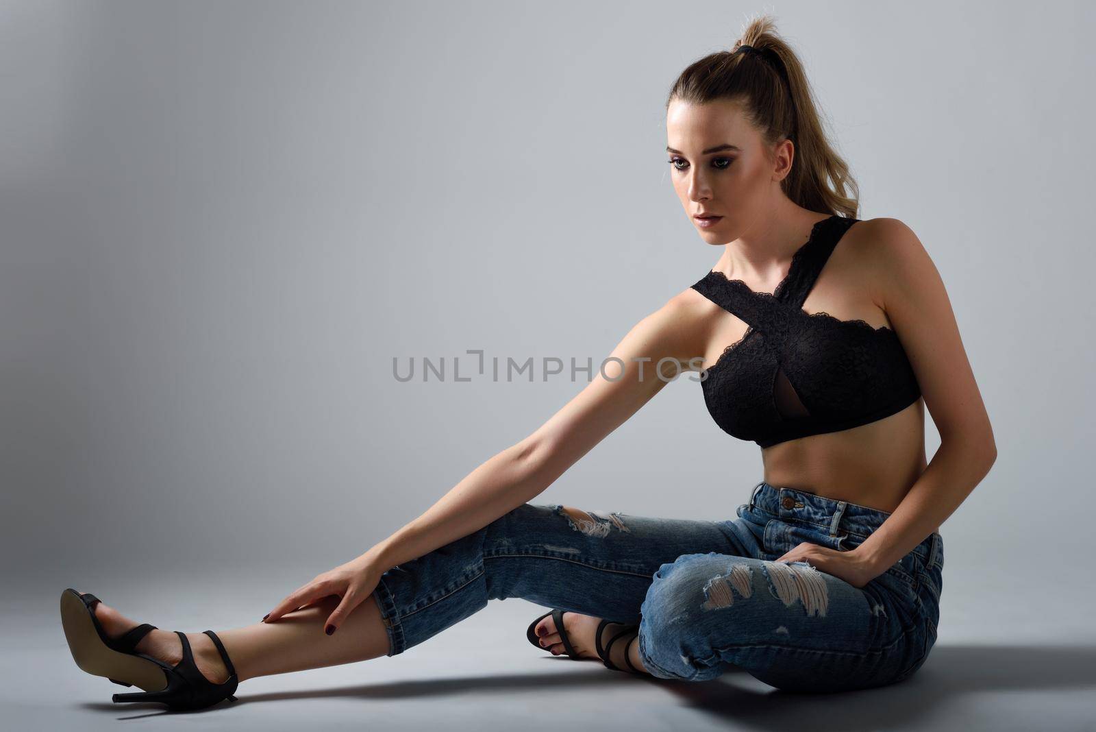 Beautiful caucasian woman with very long hair wearing black bra and blue jeans sitting on white floor. Wavy hairstyle. Studio shot.