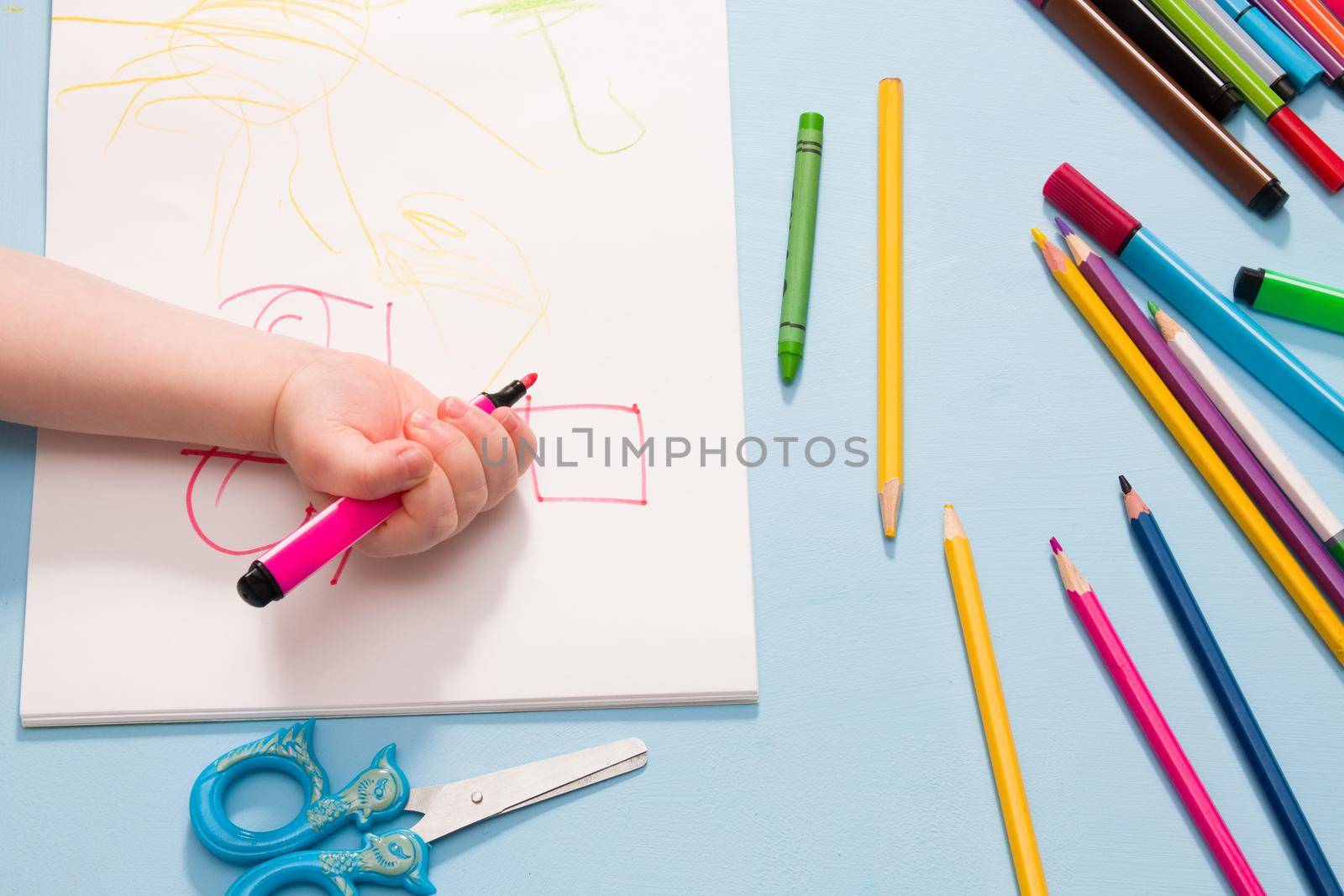 a small child draws with pencils in an album, copy space, top view, blue background, pencils, wax crayons, scissors and the child's hands on the table by natashko