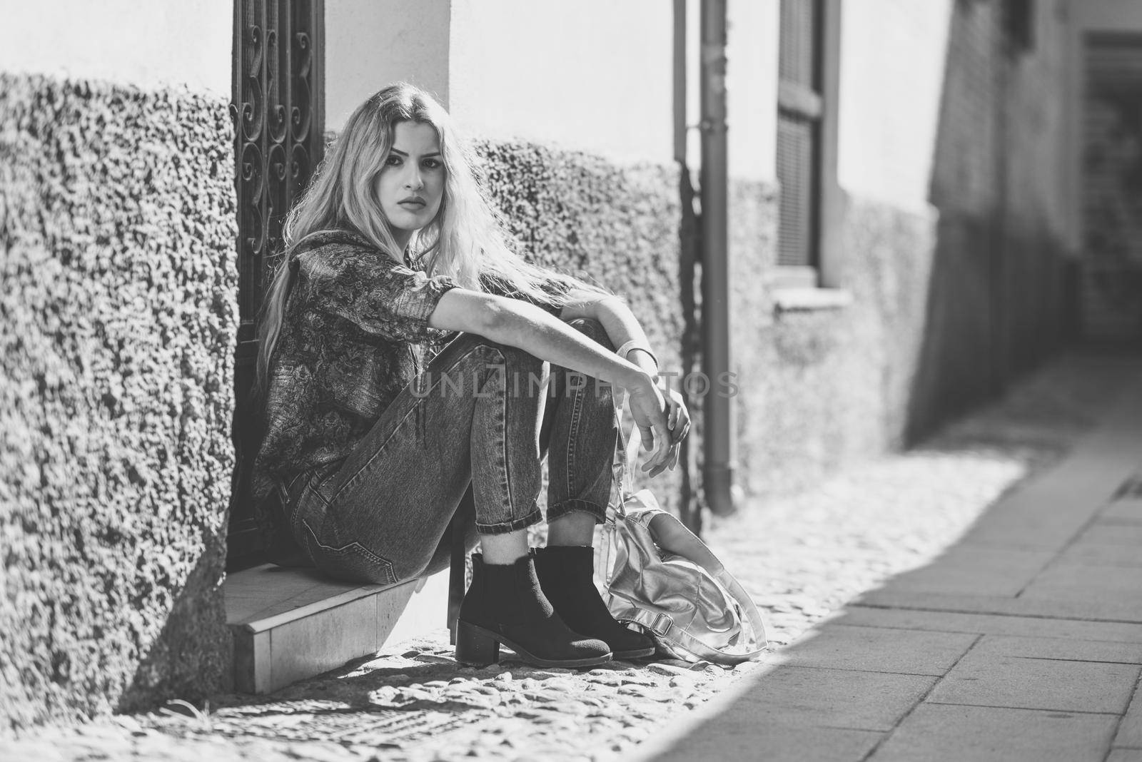 Serious blond woman, model of fashion, sitting on floor in urban background. Beautiful girl wearing shirt and blue jeans.