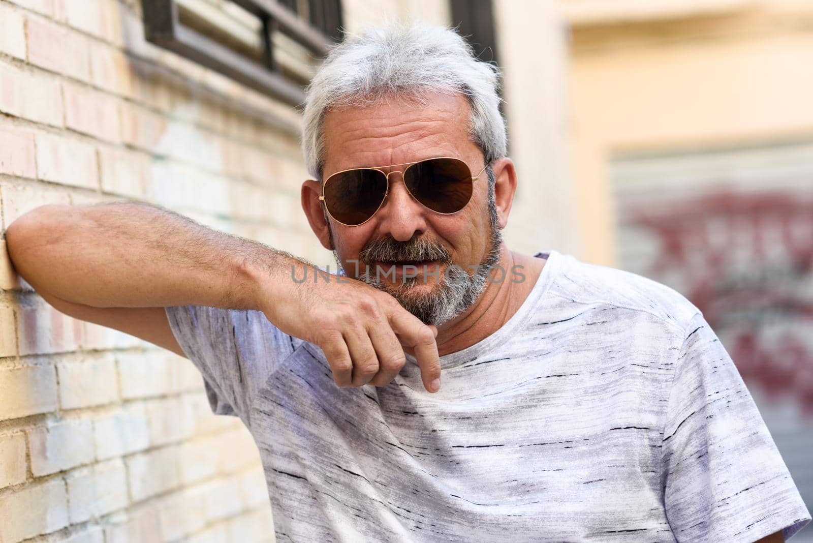 Mature man smiling at camera in urban background. Senior male with white hair and beard wearing casual clothes and aviator sunglasses.