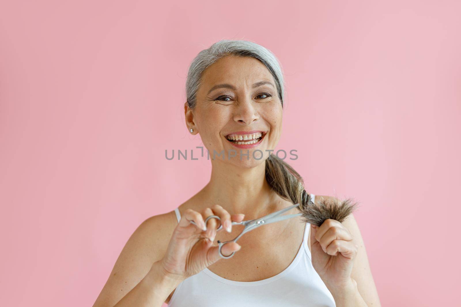 Smiling middle aged Asian lady cuts frayed ends of silver hair with scissors on pink background by Yaroslav_astakhov
