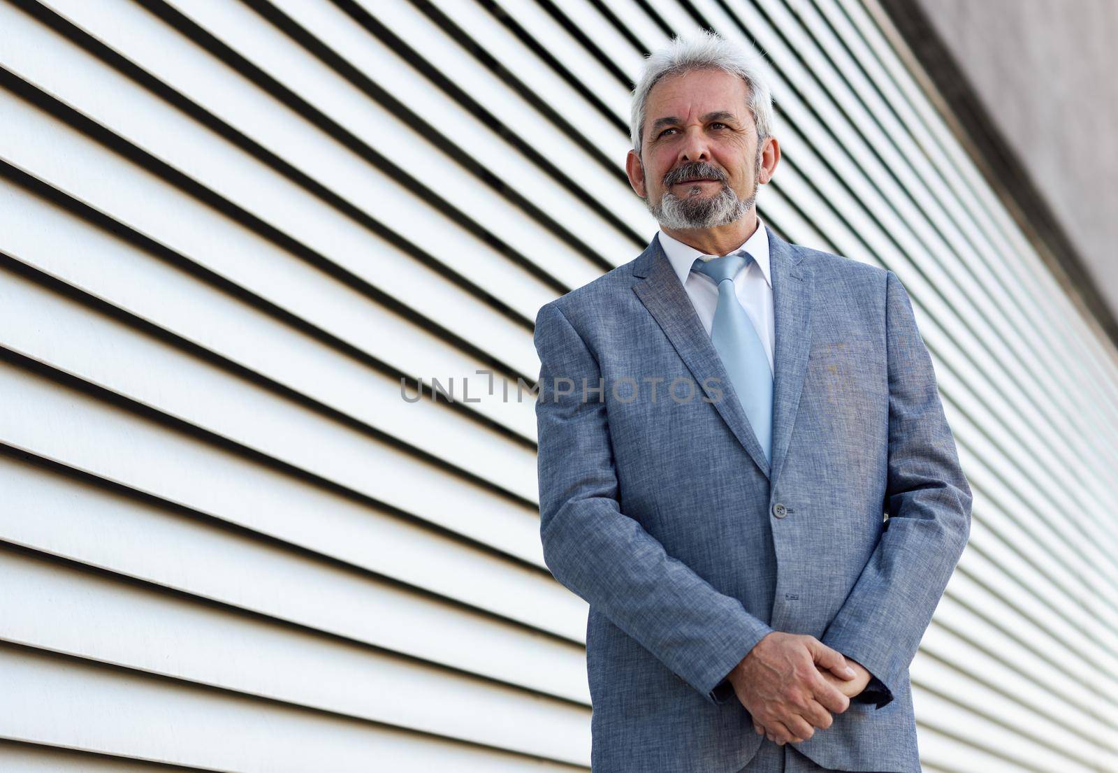 Portrait of a senior businessman with arms crossed outside of modern office building. Successful business man wearing formal suit and tie smiling in urban background.