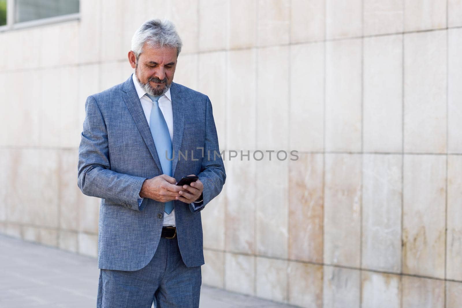 Portrait of a senior businessman texting with a smart phone outside of modern office building. Successful business man in urban background.