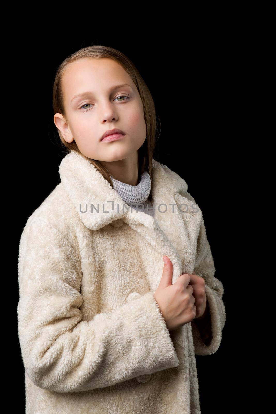 Close up portrait of stylish preteen girl. Beautiful fashionable blonde girl wearing beige faux fur coat posing against black background. Trendy child dressed fashion outfit
